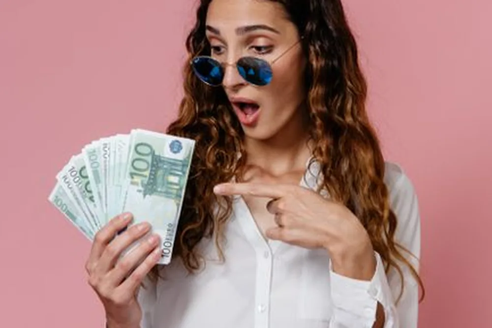 Une femme portant des lunettes de soleil tient et pointe des billets en euros, sur un fond rose.
