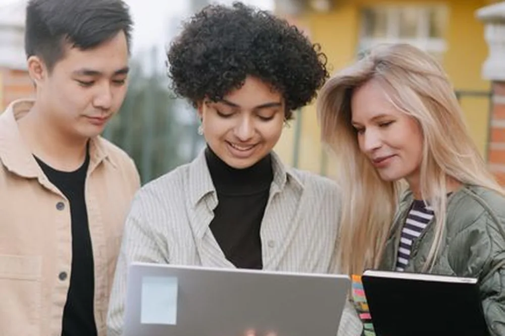 Trois jeunes adultes divers travaillant ensemble avec un ordinateur portable en plein air, symbolisant le travail d'équipe et l'intégration technologique.