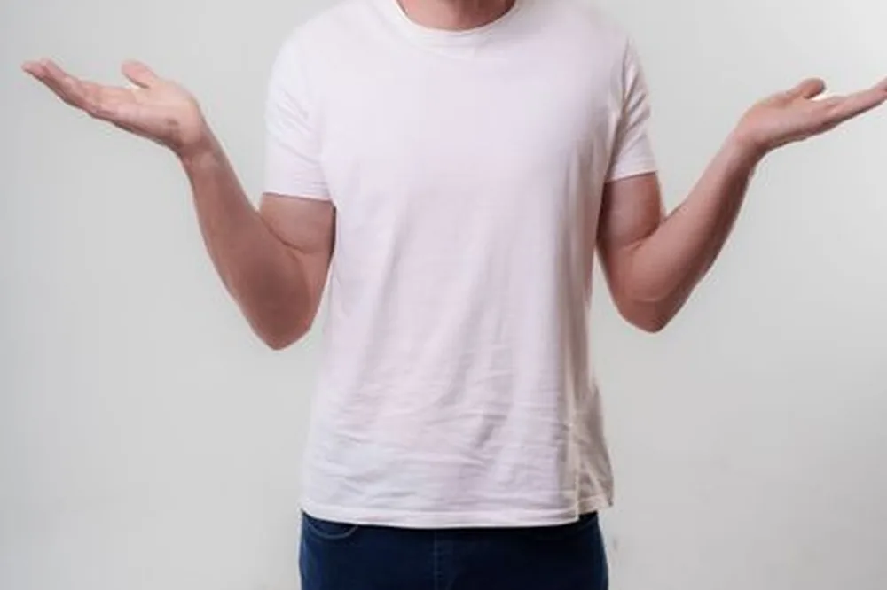 Un jeune homme adulte en vêtements décontractés hausse les épaules avec une expression neutre dans un décor de studio.